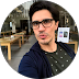 Man with dark hair and glasses wearing a dark blue jacket and green shirt, sitting indoors with tables and chairs in the background.